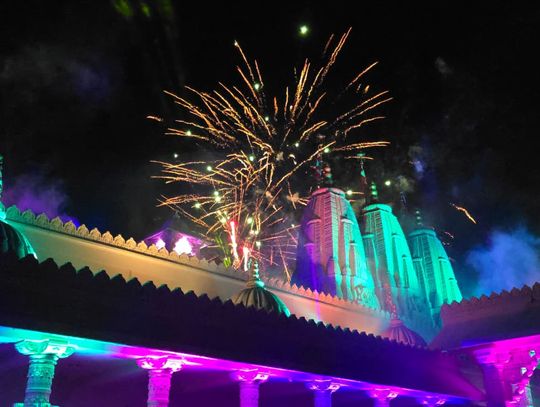 A nighttime shot of the BAPS mandir beautifully lit in vivid colors—greens, purples, yellows—while fireworks burst overhead. It gives a very festive, grand, and celebratory feel, perfect for highlighting the 25th anniversary milestone.