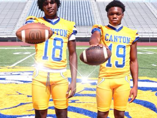 two boys in yellow canton football uniforms standing on football field holding footballs in front of them.