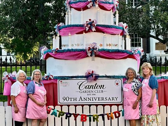 Women in pink aprons stand beside a large decorated cake display sign marking the Canton Garden Club 90th anniversary