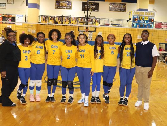 A volleyball team in yellow and blue uniforms stands in a row on a gym court with two adults