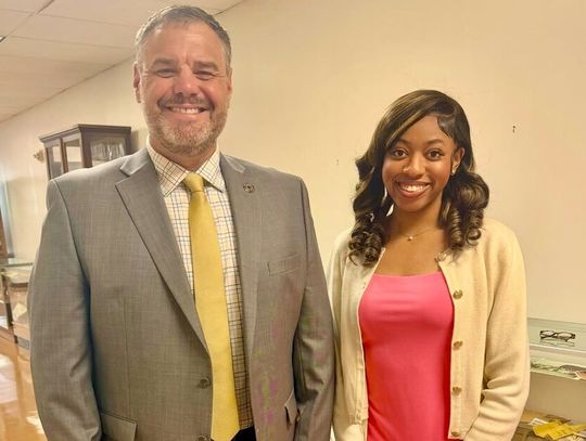 A smiling man in a gray jacket and yellow tie stands beside a smiling young woman in a pink top