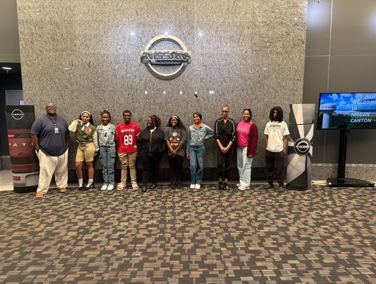 A group of students and staff stand in a row inside a large room during a visit to Nissan Canton