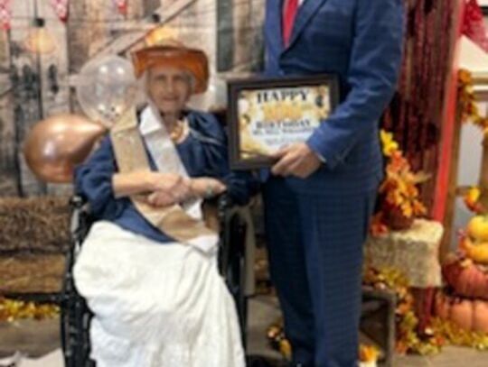 A woman in a wide-brimmed hat sits in a wheelchair beside a man in a blue suit holding a framed certificate, in front of a western-themed backdrop with sunflowers