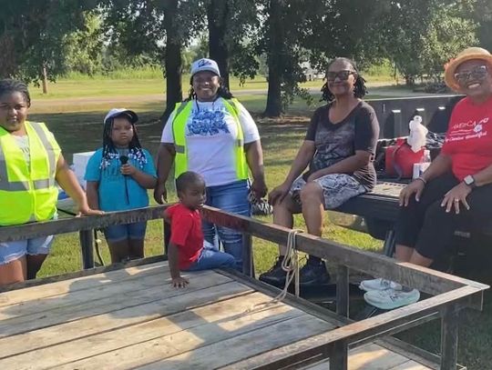 Two children and three women in safety vests sit and stand around a flatbed trailer in a park