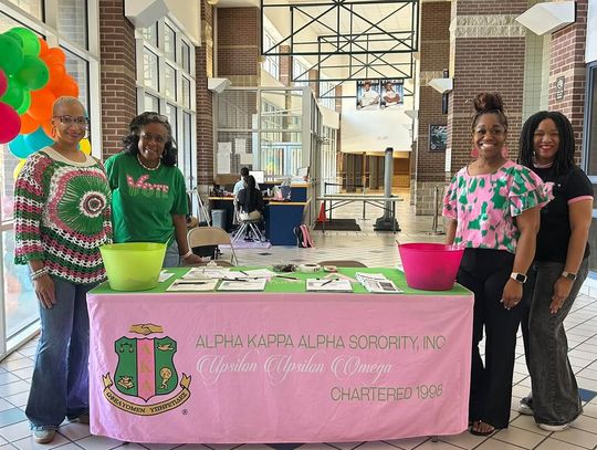 Women in pink and green stand behind a pink table with an Alpha Kappa Alpha banner and colorful balloons in a school lobby