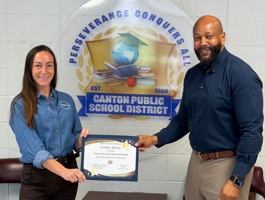 A woman and a man hold a framed certificate together in front of a Canton Public School District banner