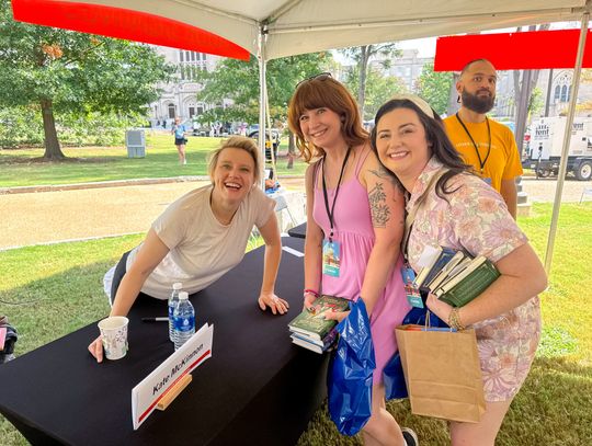 Kate McKinnon smiles behind a signing table beside two women holding books and a bag, under a tent outdoors