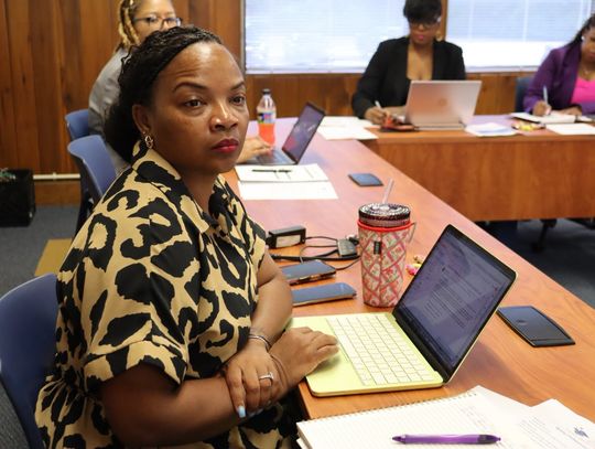 A woman in a leopard-print top sits at a conference table with a laptop and notepad while other attendees sit behind her