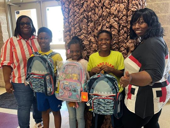 Two women stand with three students holding backpacks in front of a patterned wall