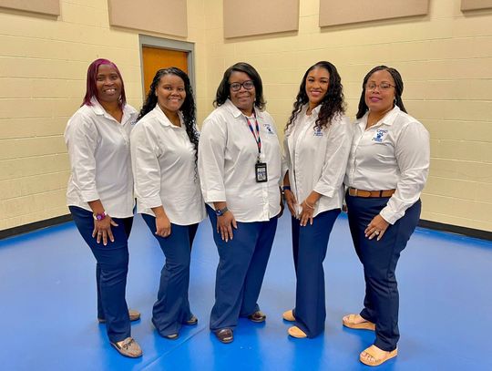 Five women in matching white shirts and navy pants stand together in a gym