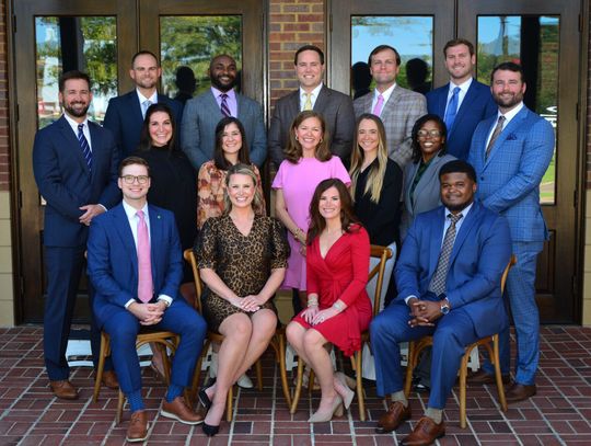 The Madison County Young Professionals board members, most in suits, pose in three rows in front of a brick wall and large windows