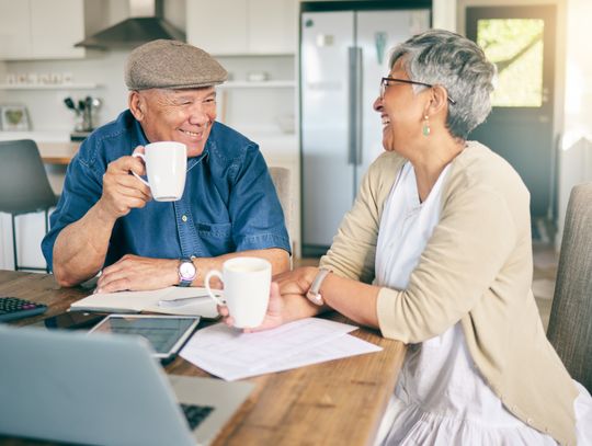 elderly couple in retirement sitting at a kitchen table laughing and talking with coffee in their hand, laptop on the table and paperwork spreadout