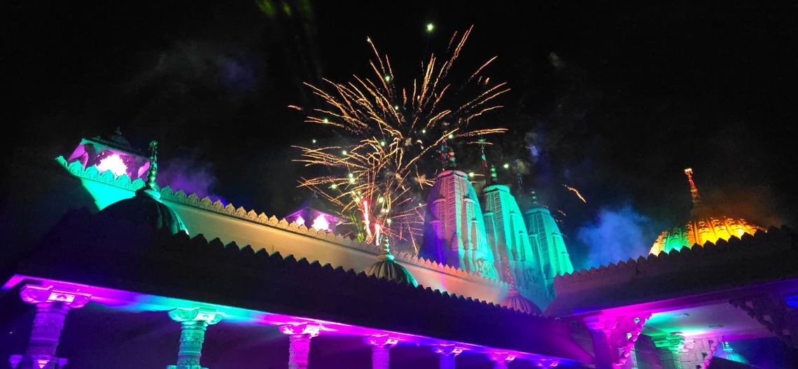 A nighttime shot of the BAPS mandir beautifully lit in vivid colors—greens, purples, yellows—while fireworks burst overhead. It gives a very festive, grand, and celebratory feel, perfect for highlighting the 25th anniversary milestone.