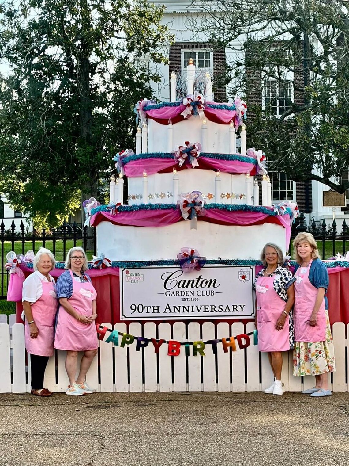 Canton Garden Club celebrates 90 years of community beautification Women in pink aprons stand beside a large decorated cake display sign marking the Canton Garden Club 90th anniversary