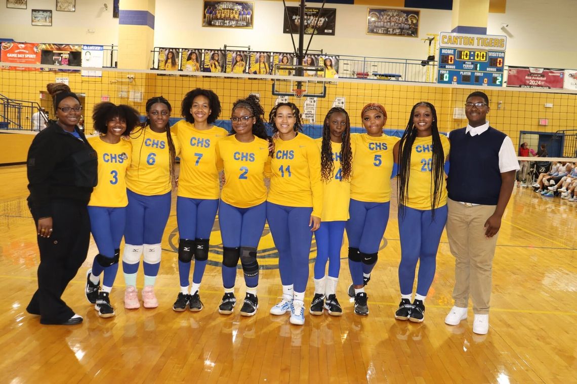 A volleyball team in yellow and blue uniforms stands in a row on a gym court with two adults