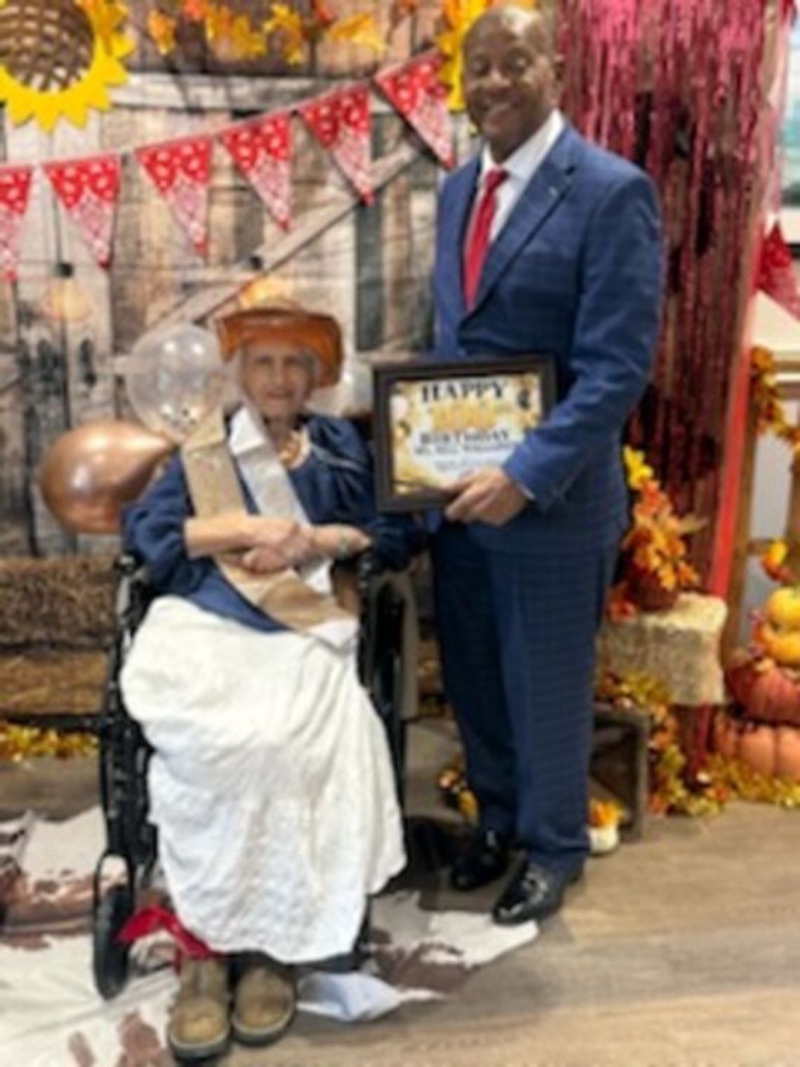 Canton woman celebrates 100th birthday A woman in a wide-brimmed hat sits in a wheelchair beside a man in a blue suit holding a framed certificate, in front of a western-themed backdrop with sunflowers