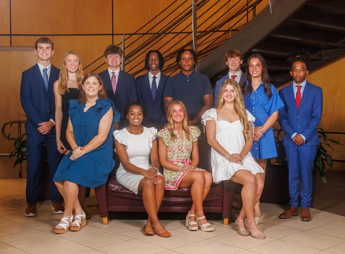 Madison County Young Professionals announces 2026-27 board Twelve young people in formal clothes pose together, some standing and some seated on a bench, in front of a staircase