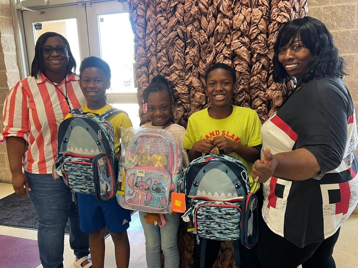 Two women stand with three students holding backpacks in front of a patterned wall