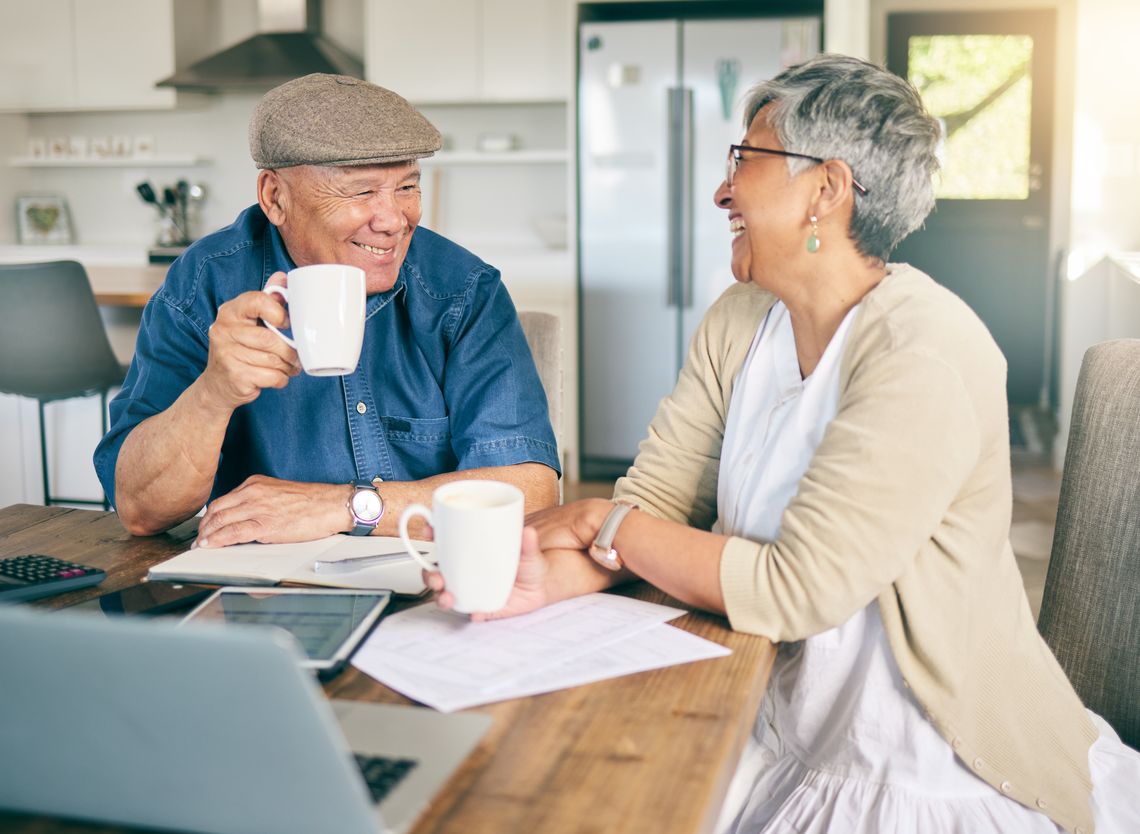 elderly couple in retirement sitting at a kitchen table laughing and talking with coffee in their hand, laptop on the table and paperwork spreadout