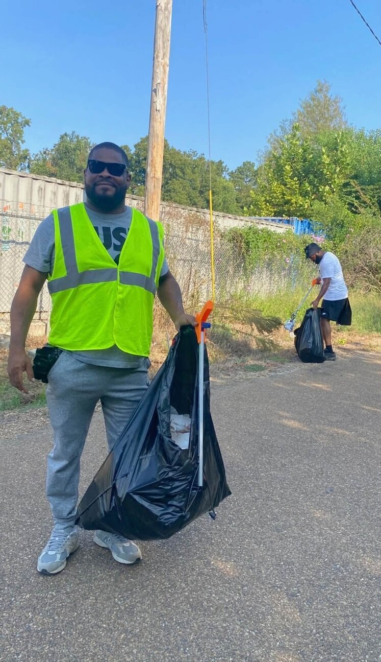 A volunteer in a safety vest holds a full black trash bag and a litter grabber beside a fence while another volunteer picks up litter behind him
