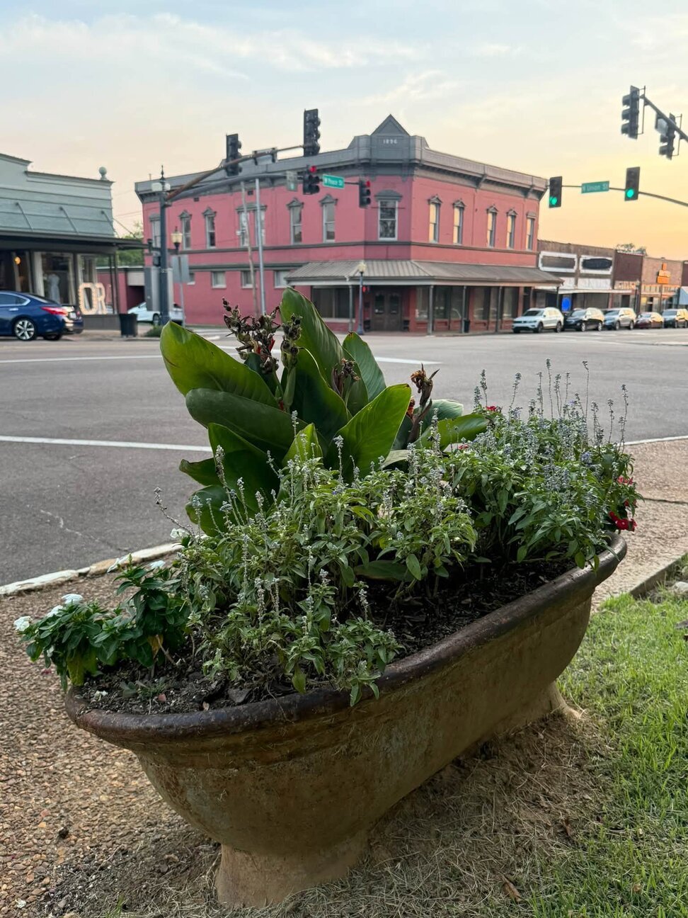 A stone trough planter filled with green plants and flowers on a downtown street corner with a red brick building behind it