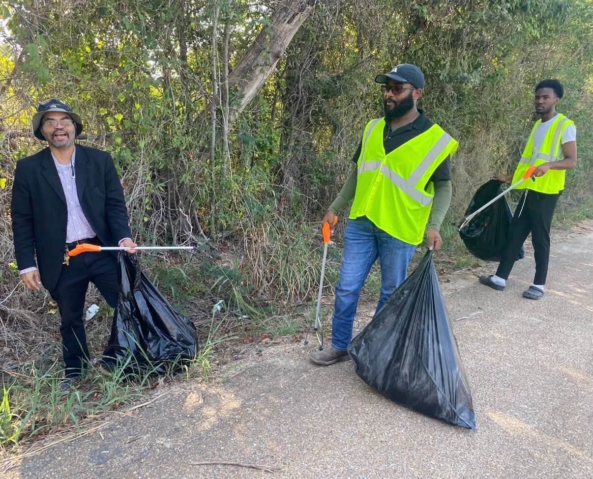 Three volunteers pick up litter with grabbers and trash bags along a weedy roadside