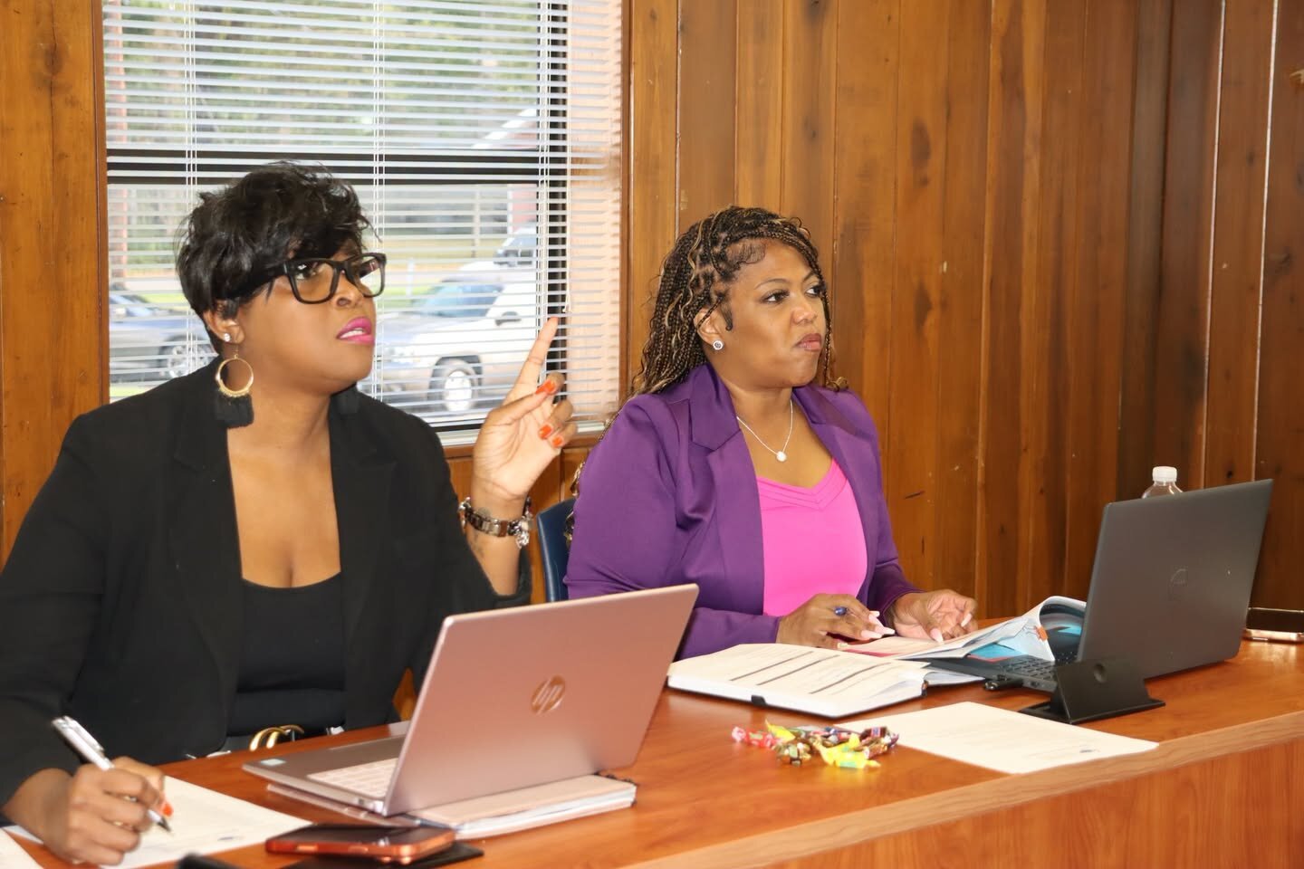 Two women sit side by side at a desk with laptops and notebooks, one speaking and gesturing