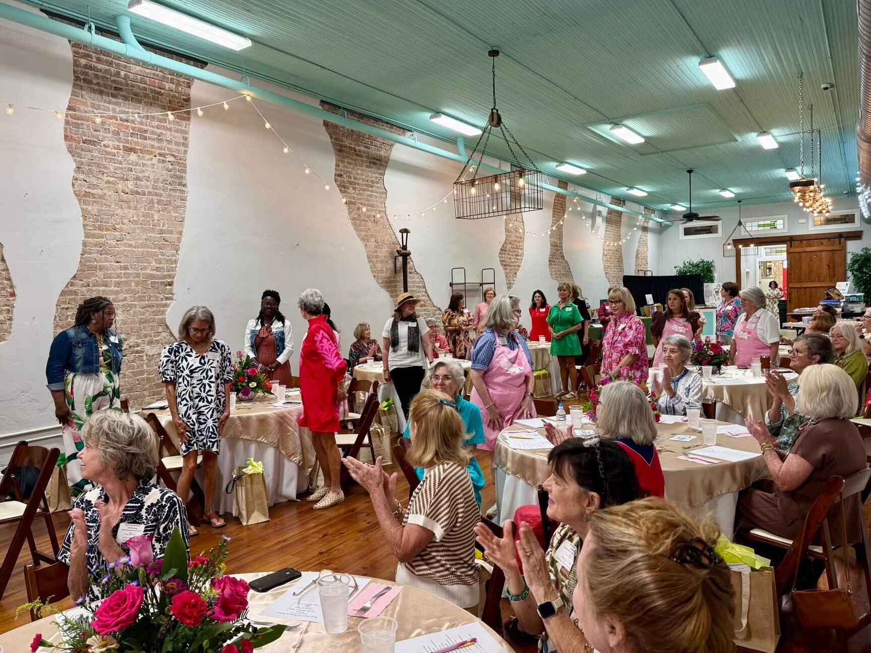 Guests in dresses gather at round tables in a large brick-walled event hall during the Canton Garden Club celebration