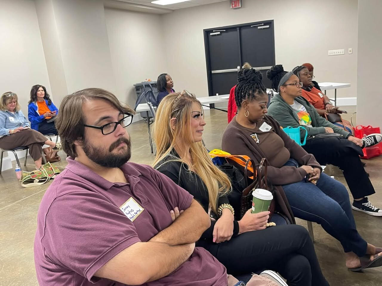 A man in a purple shirt and a woman with long blond hair sit in a room full of conference attendees