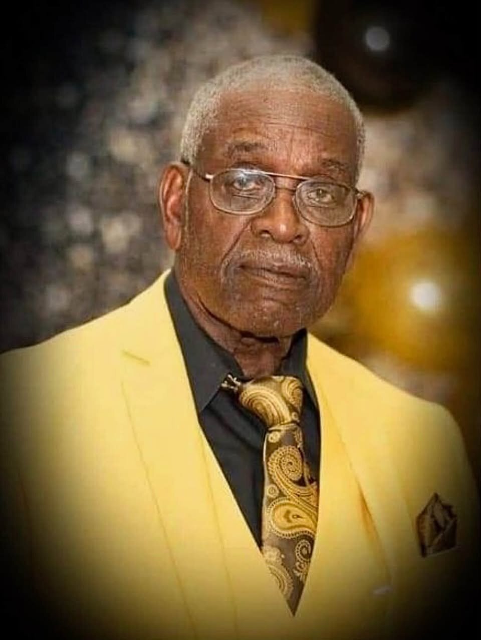 Portrait of an older man with glasses wearing a yellow jacket and a gold patterned tie