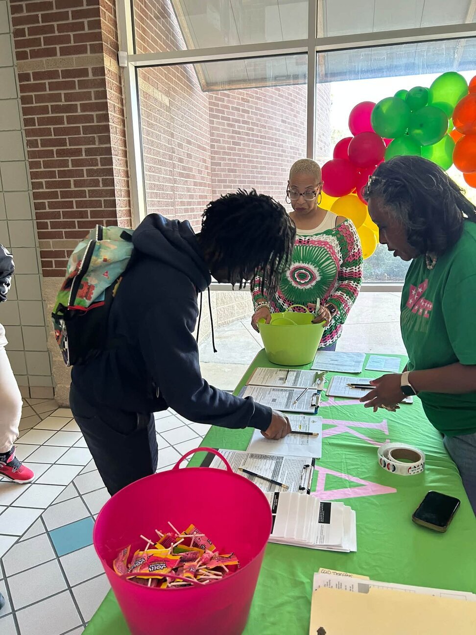A student leans over a green table to fill out paperwork while two women stand nearby, with a pink bucket in the foreground