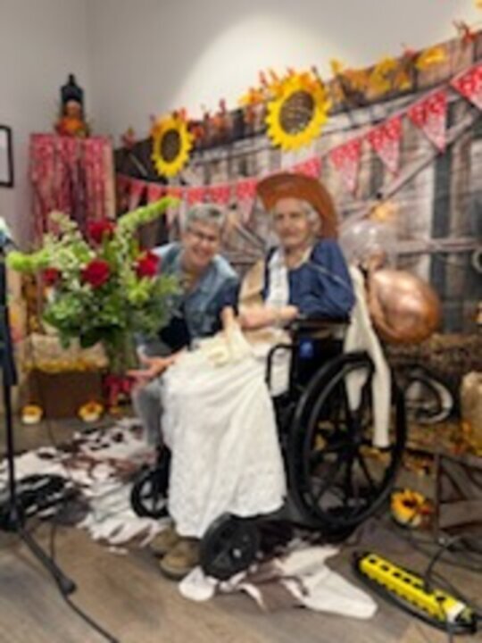 Two women sit near a bouquet of red roses in front of a western-themed backdrop decorated with sunflowers