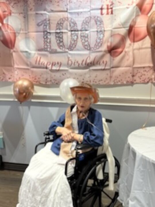 A woman in a straw hat sits in a wheelchair in front of a pink 100th Happy Birthday banner